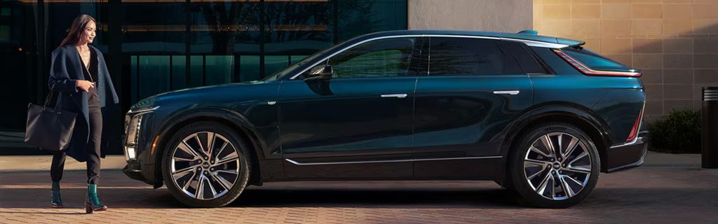 Woman walking toward a sleek dark green 2026 Cadillac LYRIQ parked outside a modern building.